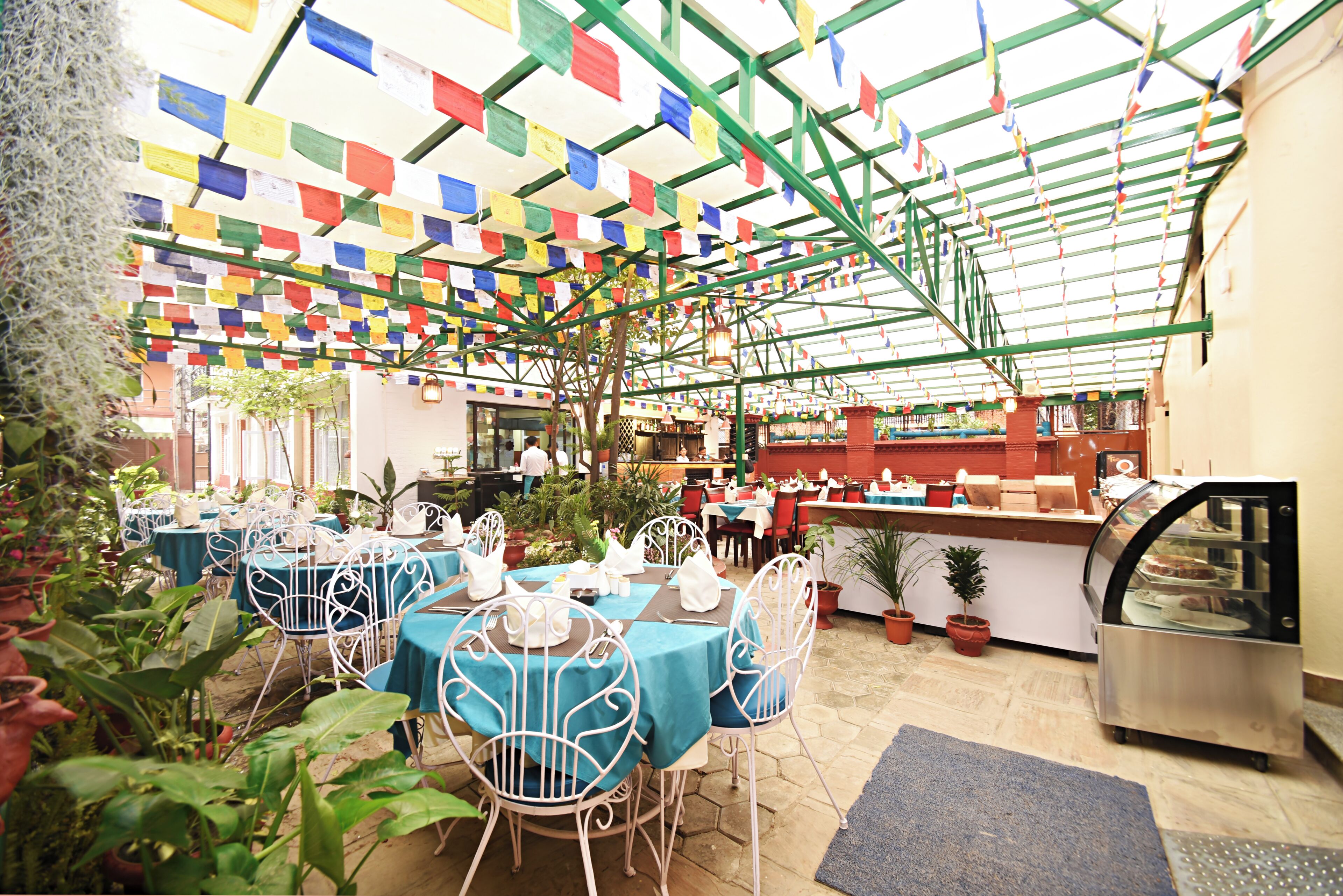 Ein Restaurant mit Tischen, die mit blauen Tischdecken bedeckt sind, weißen Stühlen, einer Glasvitrine und einer grünen Metallkonstruktion darüber.