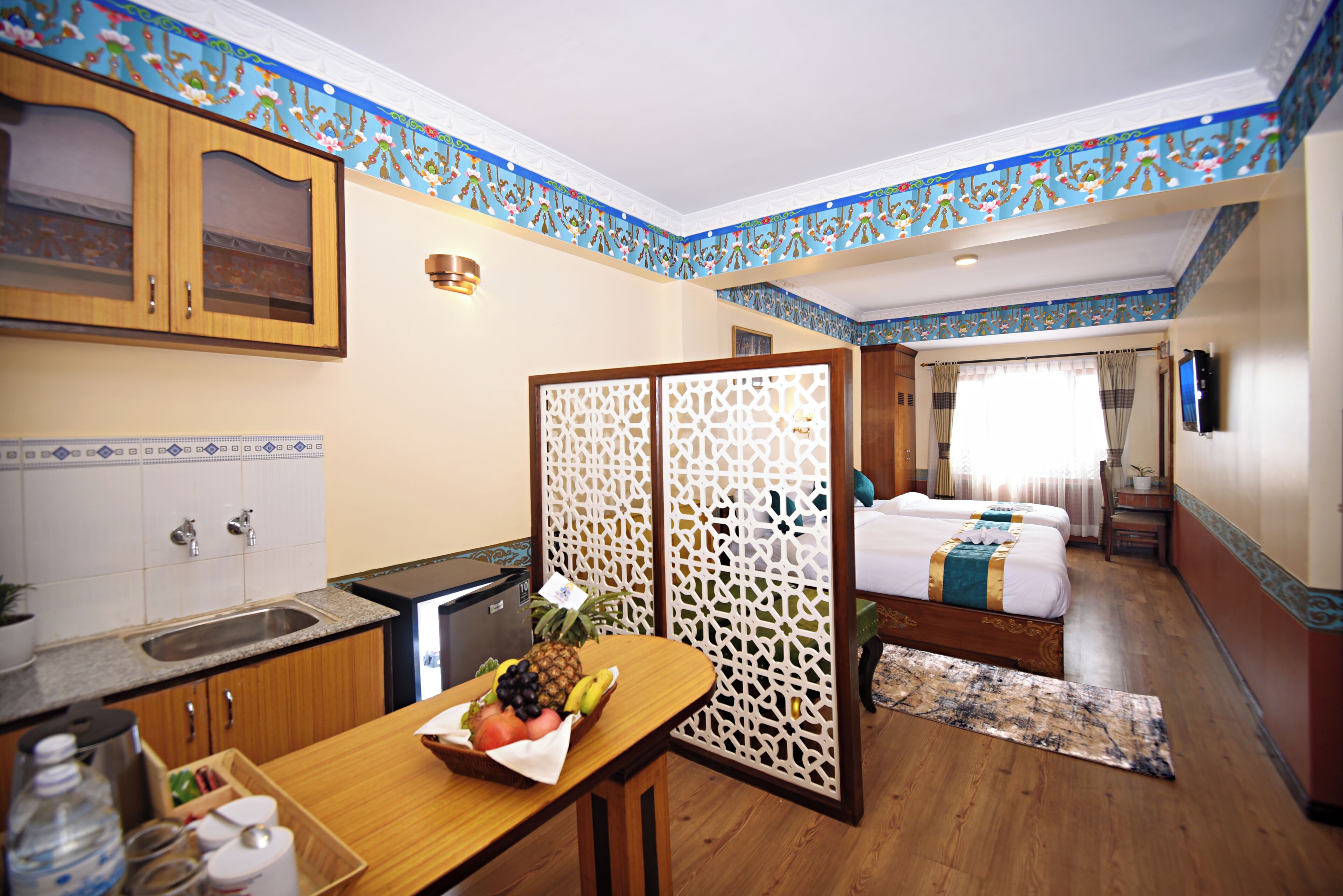 A hotel room with a kitchen area, a dining table with fruit, a bed, and a TV.