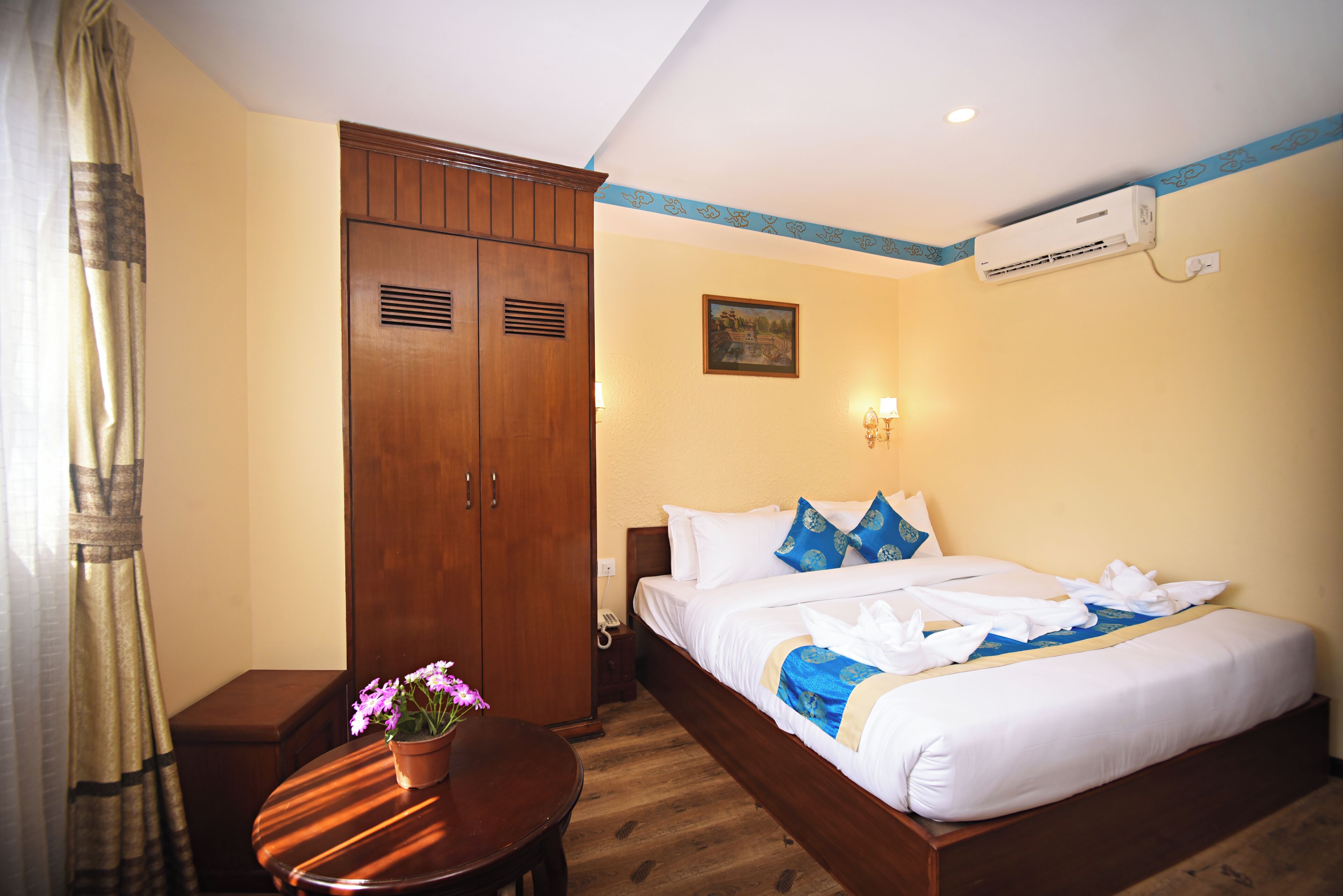 Habitación de hotel con un armario de madera, cama con ropa de cama blanca y detalles azules, una mesita redonda con una planta en maceta y un equipo de aire acondicionado en la pared.