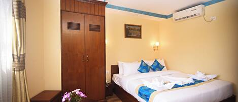Habitación de hotel con un armario de madera, cama con ropa de cama blanca y detalles azules, una mesita redonda con una planta en maceta y un equipo de aire acondicionado en la pared.