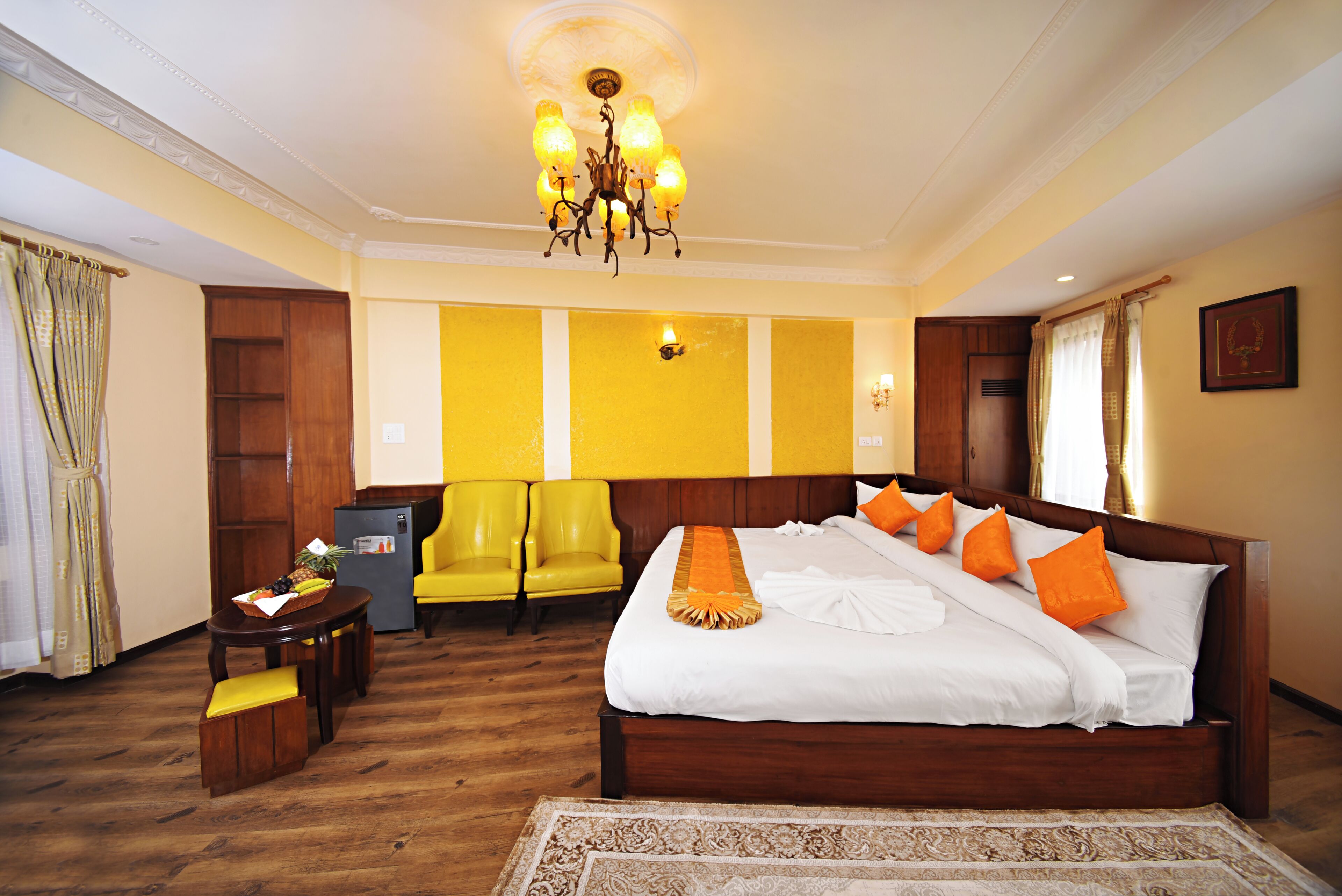 A hotel room with a large bed, a wooden headboard, a chandelier, a desk with a chair, and a small table with a fruit bowl.