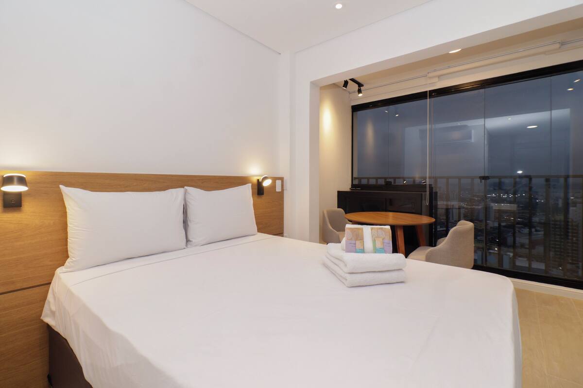 Habitación de hotel con una cama grande, una mesita redonda y vistas a la ciudad a través de un ventanal amplio.