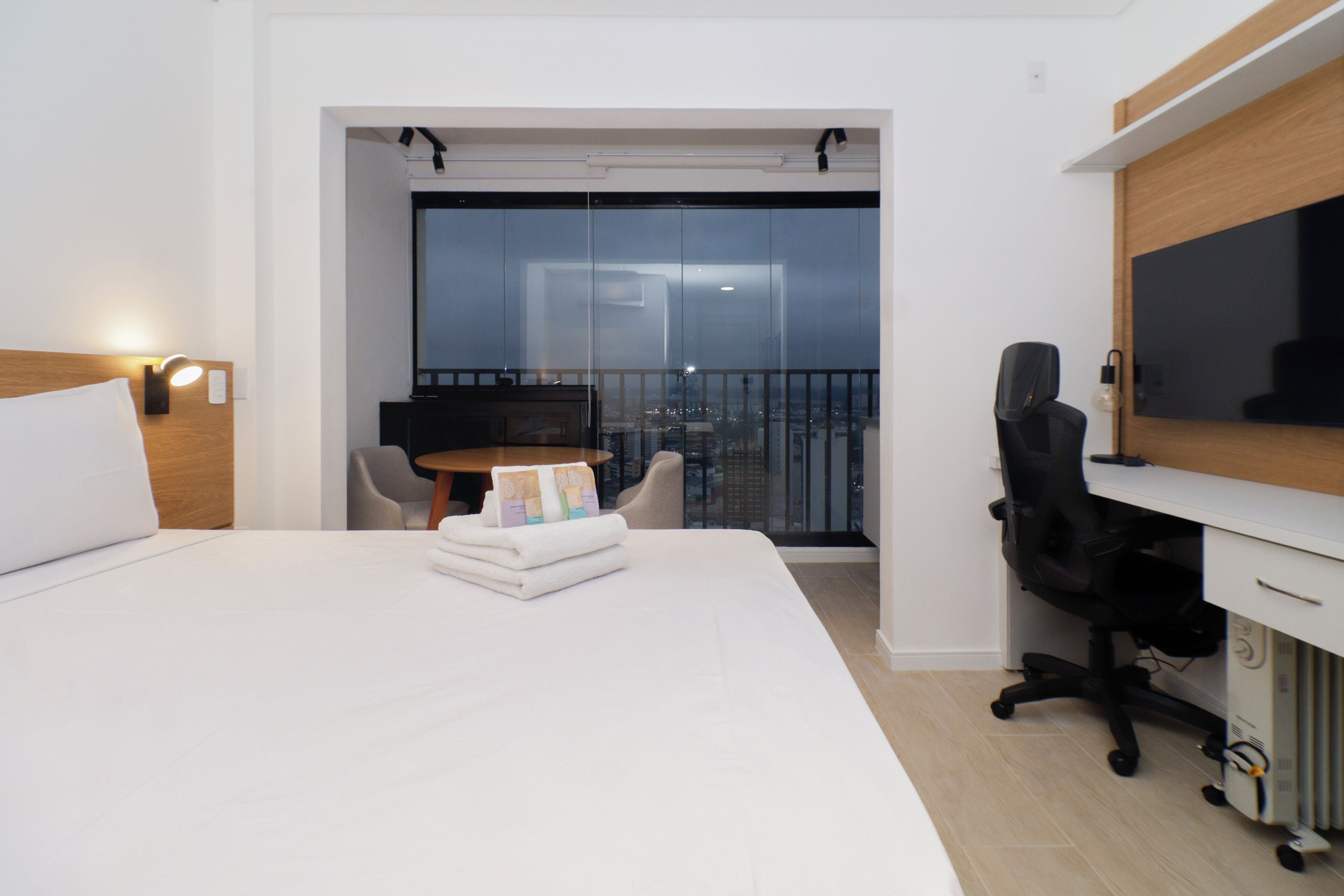 A hotel room with a bed, a desk with a computer, a chair, and a view of the city through a glass door.