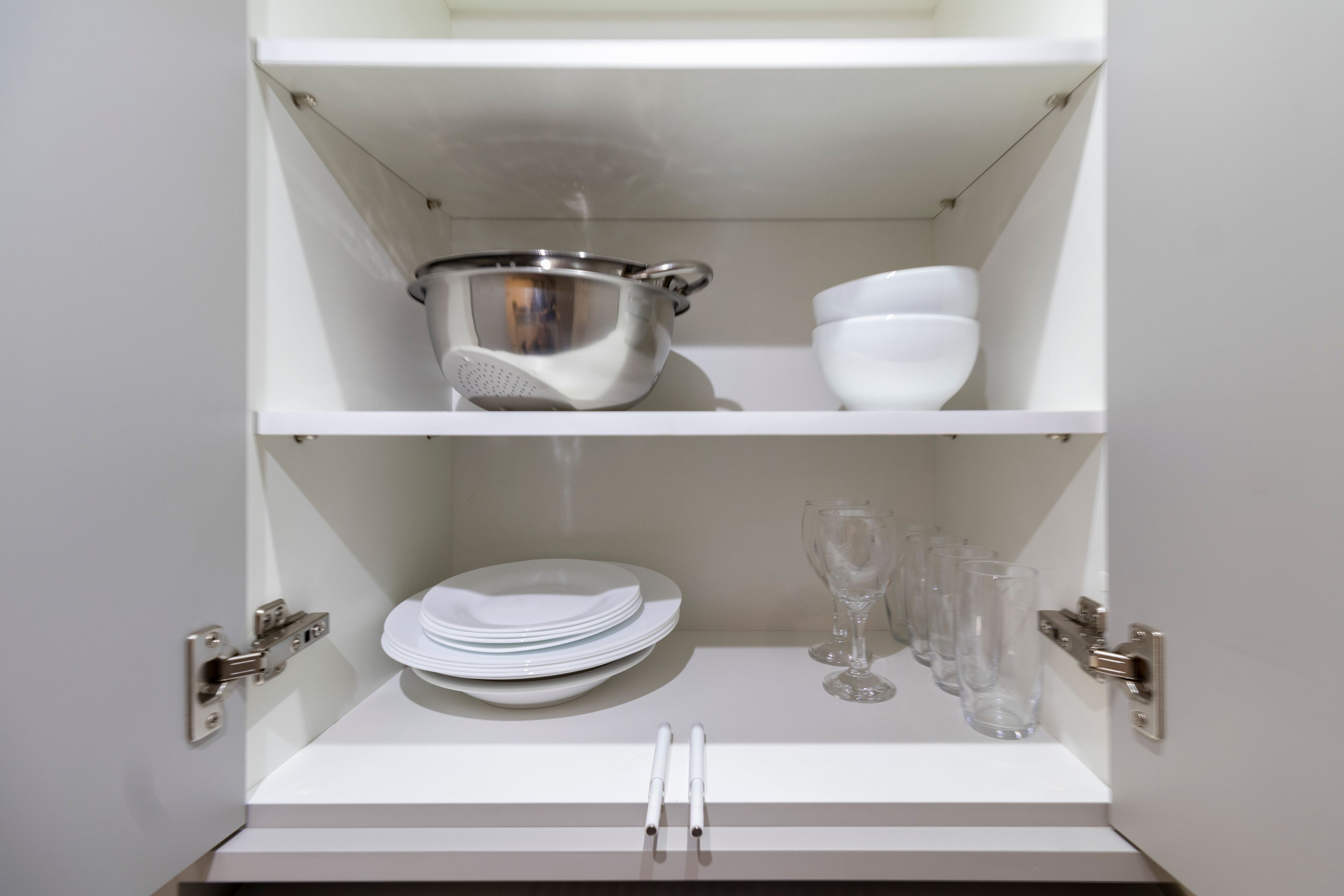 Un espace intérieur aux murs blancs, avec une étagère contenant une passoire en acier inoxydable, une pile d’assiettes blanches et plusieurs verres transparents.