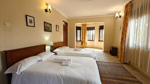 A hotel room with two beds, a wooden headboard, a small table with a lamp, and a colorful rug. - Entebbe Coffee Lodge (Entebbe)