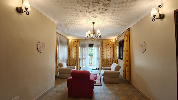 A hallway with a red armchair, beige armchairs, a chandelier, patterned curtains, and a ceiling with a textured design. - Entebbe Coffee Lodge (Entebbe)
