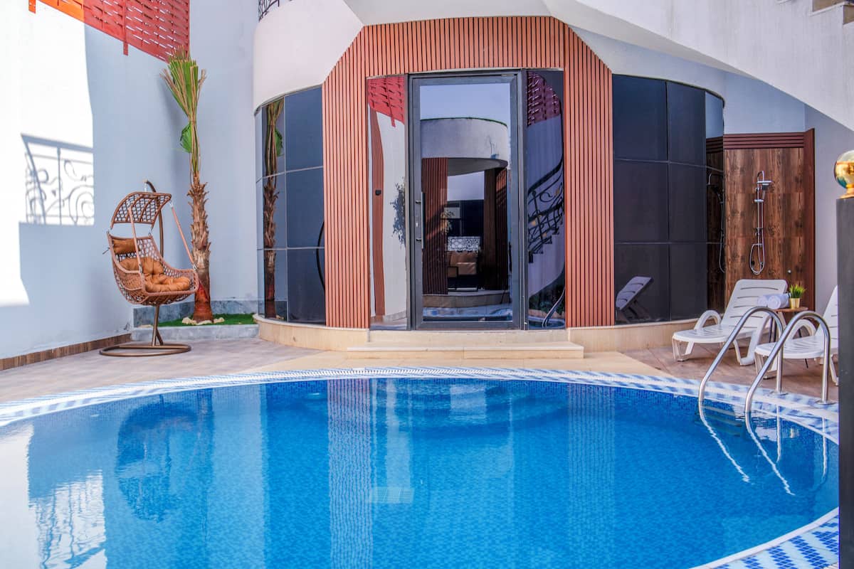 A modern indoor swimming pool with clear blue water, surrounded by lounge chairs and a wooden structure with glass doors.