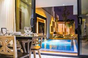 A dining area with a pool view, featuring a table set for two, chairs, and a glass partition. - The Land Resort (Riyadh)