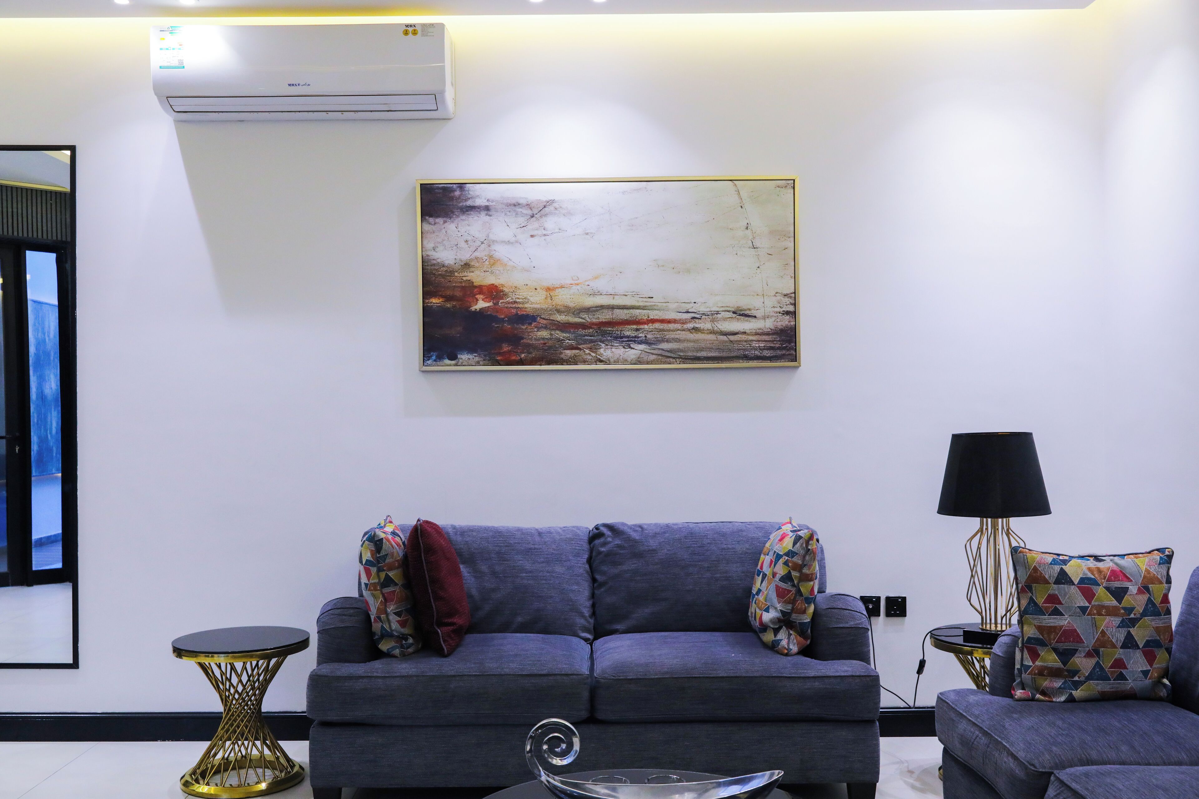 A modern living room with a grey sofa, a colorful abstract painting, and a small side table with a lamp.