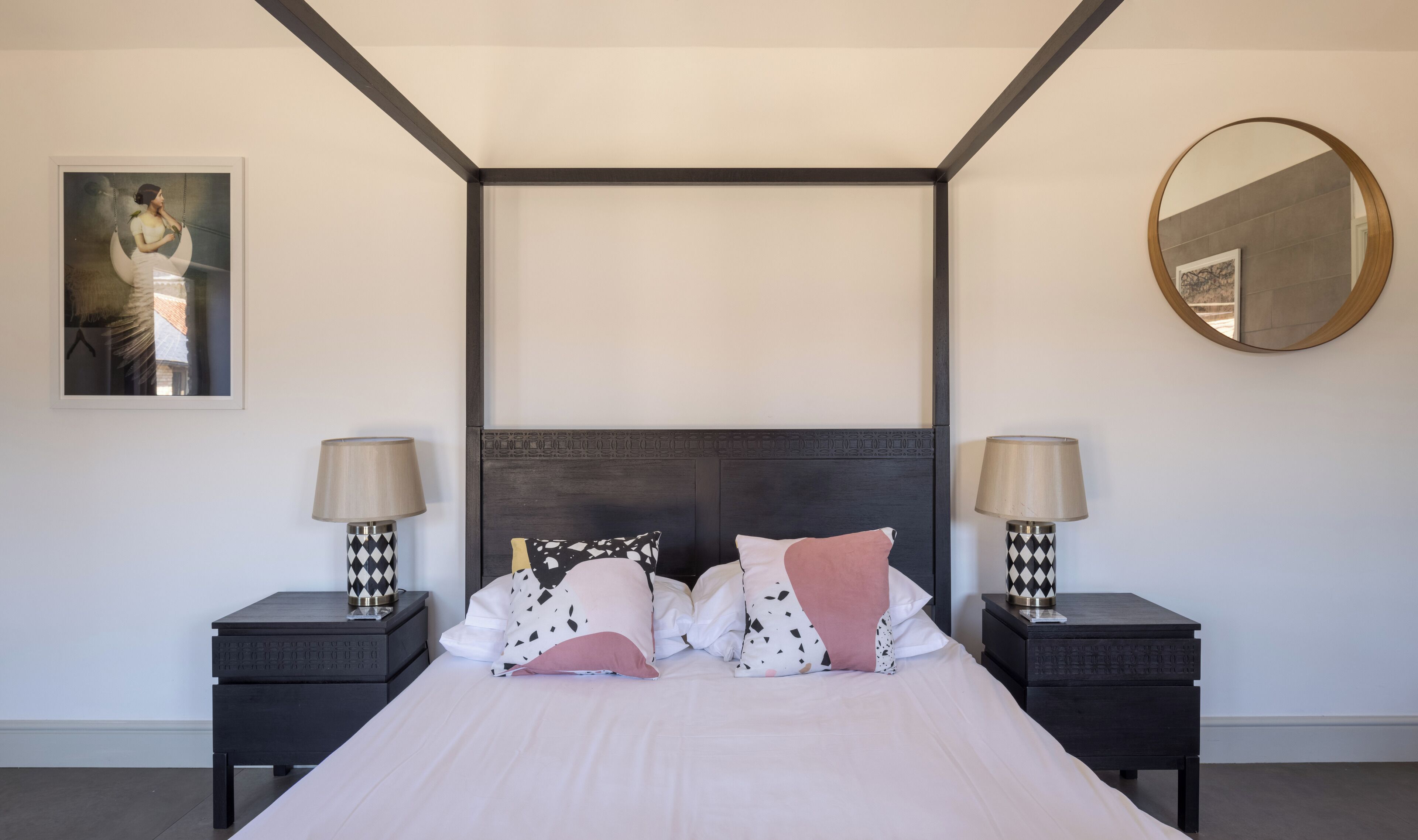 A modern bedroom with a large bed, two bedside tables with lamps, a dark headboard, a round mirror, and a framed picture on the wall.