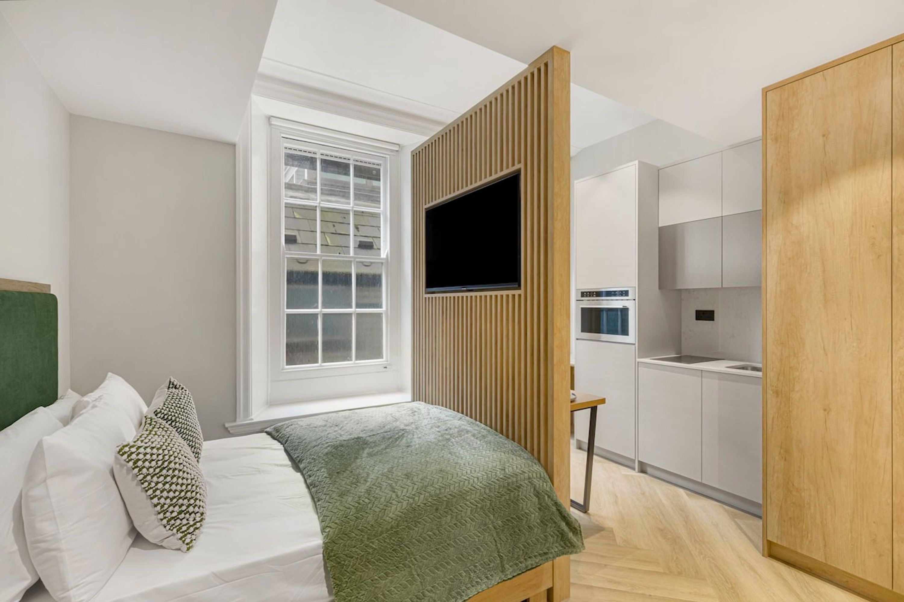 Un dormitorio moderno con cama, televisor, ventana y zona de cocina.