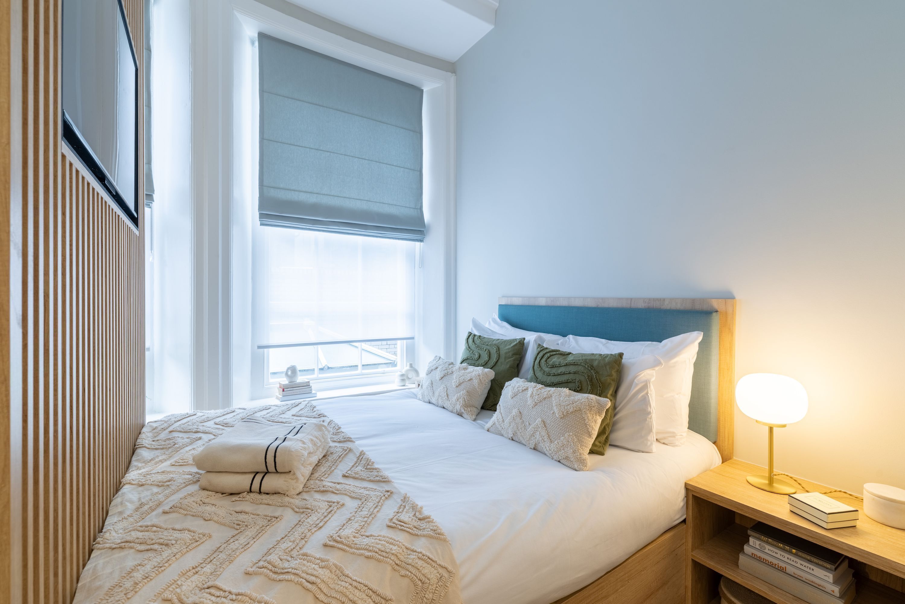 Una cama bien hecha con un edredón estampado, una mesita de noche con una lámpara y una ventana con persiana.