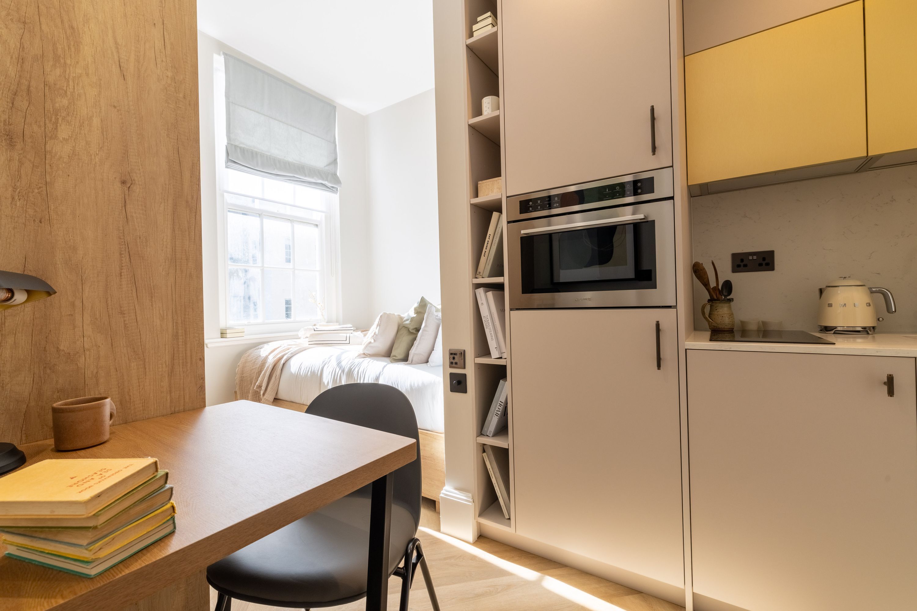 A compact bedroom with a bed, bedside table, and a window with blinds. Adjacent is a kitchenette with a microwave, oven, and a coffee maker.