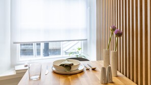 Una mesa de comedor con un tazón, vaso y cubiertos, un florero con flores, y una ventana con persianas.