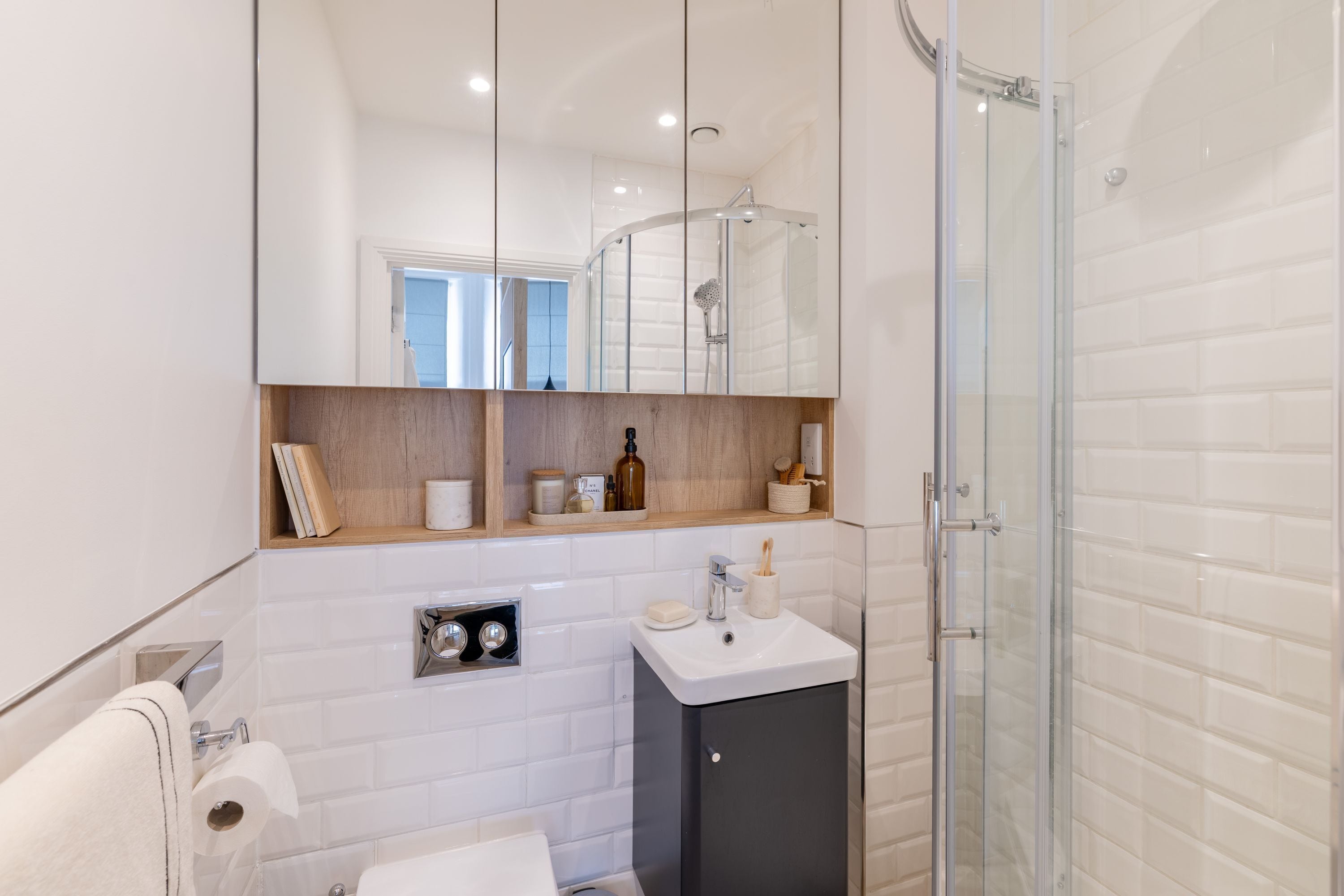 Une salle de bain moderne avec une douche à parois de verre, un lavabo blanc et une étagère en bois où sont rangés les articles de toilette.