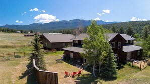 Una casa grande de madera con varias habitaciones, rodeada por un patio cercado y un paisaje montañoso de fondo.