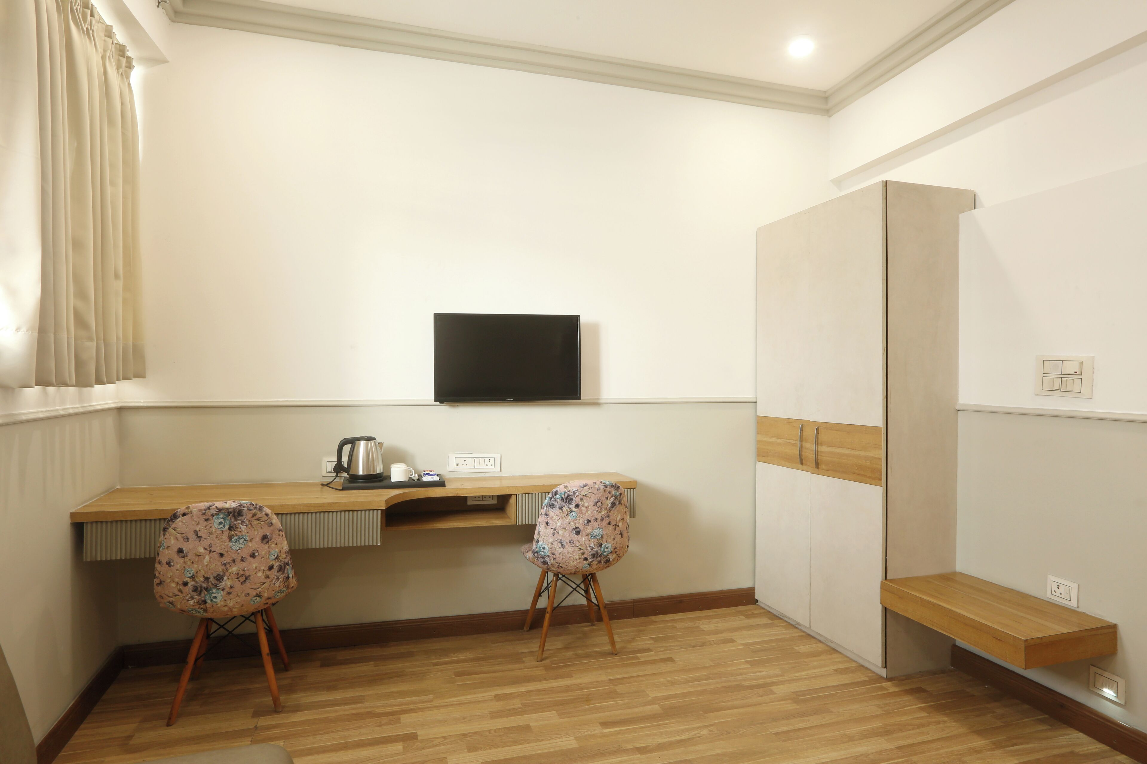 Une chambre avec un bureau en bois, deux chaises, un téléviseur à écran plat, une bouilloire et une armoire.