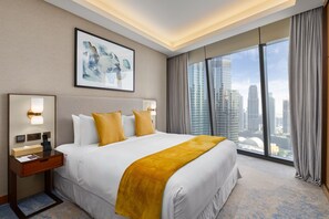 A hotel room with a large bed, a nightstand with a lamp, a framed artwork on the wall, and a city view through large windows.