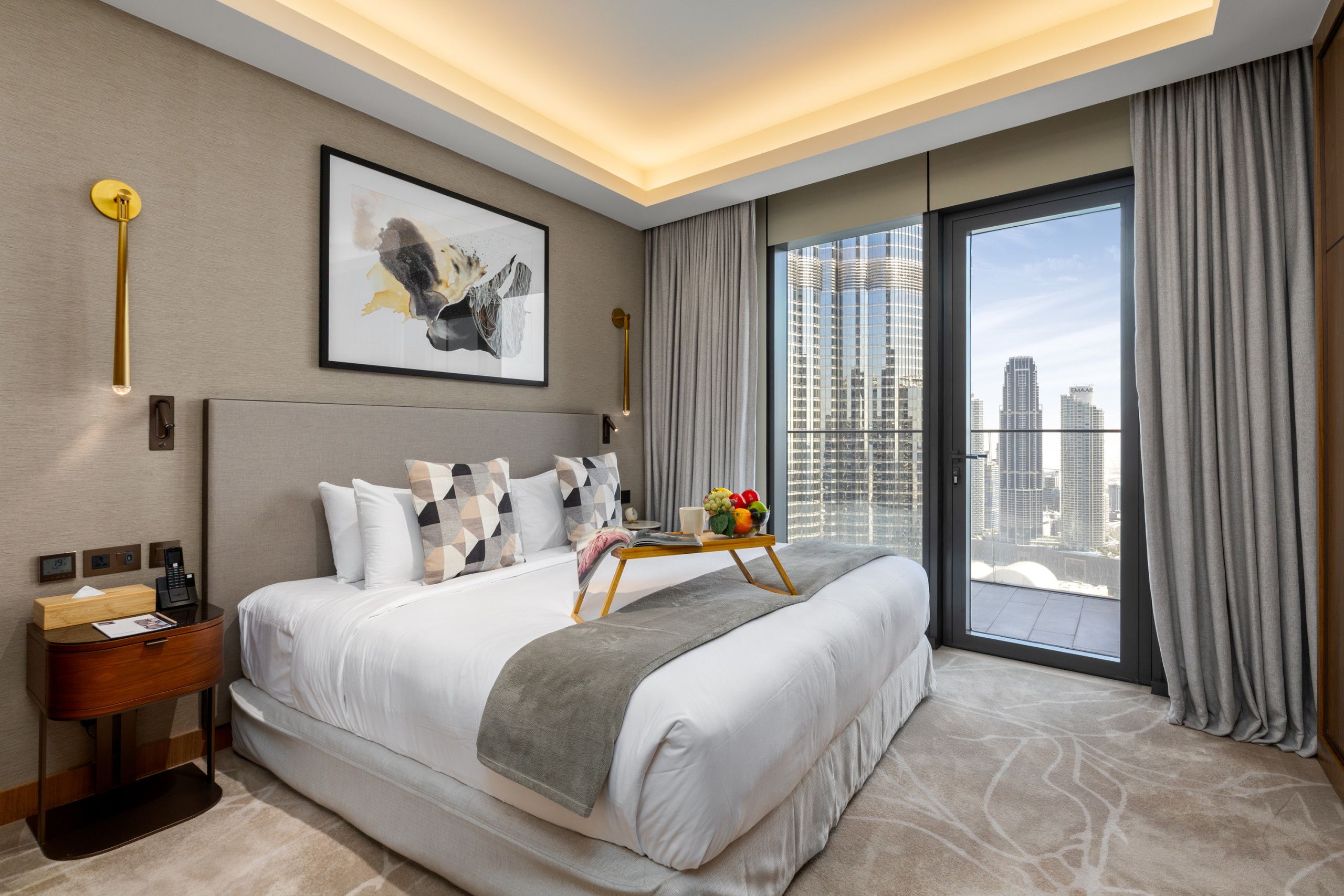 A modern hotel room with a large bed, a bedside table, a nightstand, a framed artwork, a window with a city view, and a balcony.