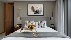 Moderni hotellihuone, jossa on suuri sänky, yöpöytä hedelmäkulholla, seinälle ripustettu abstrakti taulu ja näkymä kaupunkiin.