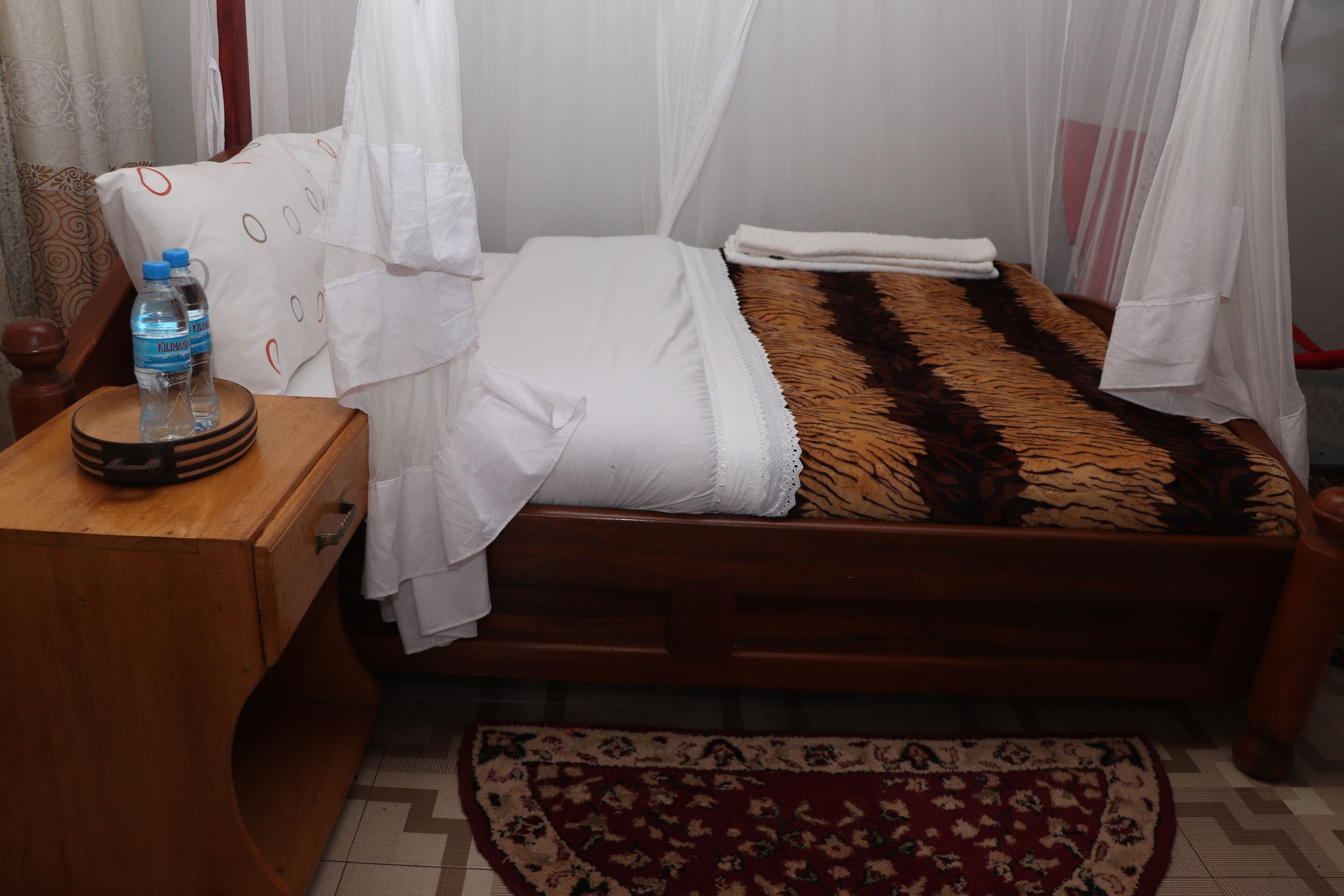 Una habitación con una cama, una mesita de noche de madera, una botella de agua y una alfombra estampada.