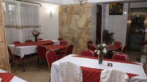 A restaurant with multiple tables, each covered with a white tablecloth and red plaid table runners. The walls are decorated with a stone pattern and a painting. - Marangu Summit Lodge (Marangu)