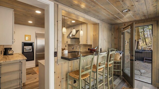 A kitchen with wooden cabinets, a stainless steel refrigerator, and a built-in oven. There are bar stools and a large window overlooking a garden. - Hottub/Arcade/Historic Cabin/Mins to AVL & WVL (Weaverville)