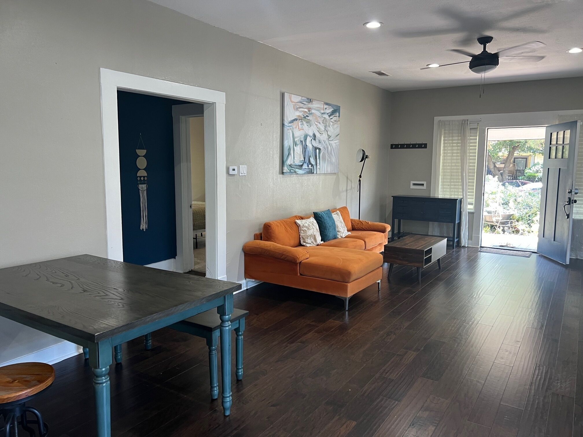 Amplia sala de estar con piso de madera oscura, una mesa azul y un sofá naranja con cojines.
