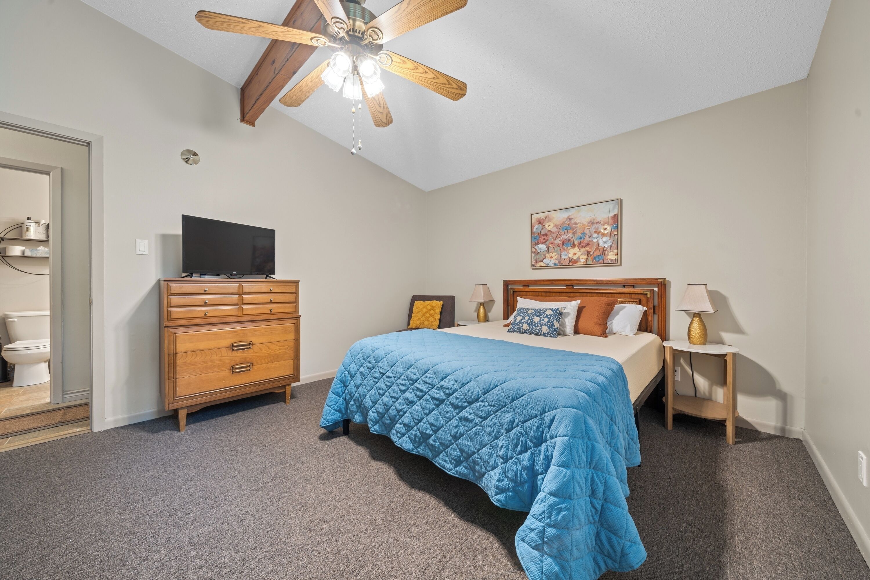 Une chambre à coucher avec un lit, une commode en bois, un ventilateur de plafond, un téléviseur et un tableau au mur.