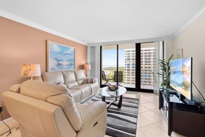 A modern living room with a beige sofa, a glass coffee table, and a flat-screen TV. There is a large window with a view of buildings and a balcony. - Sun, Sand, and Serenity I Beachfront on Marco Island (Marco Island)