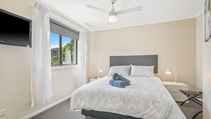 Une chambre à coucher comprenant un lit, un ventilateur de plafond, une fenêtre avec des rideaux, une table de chevet avec une lampe et un téléviseur fixé au mur.