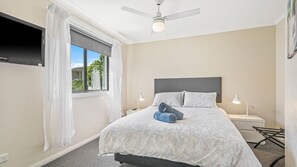 Een slaapkamer met een bed, een plafondventilator, een raam met gordijnen, een nachtkastje met een lamp en een aan de muur bevestigde televisie.