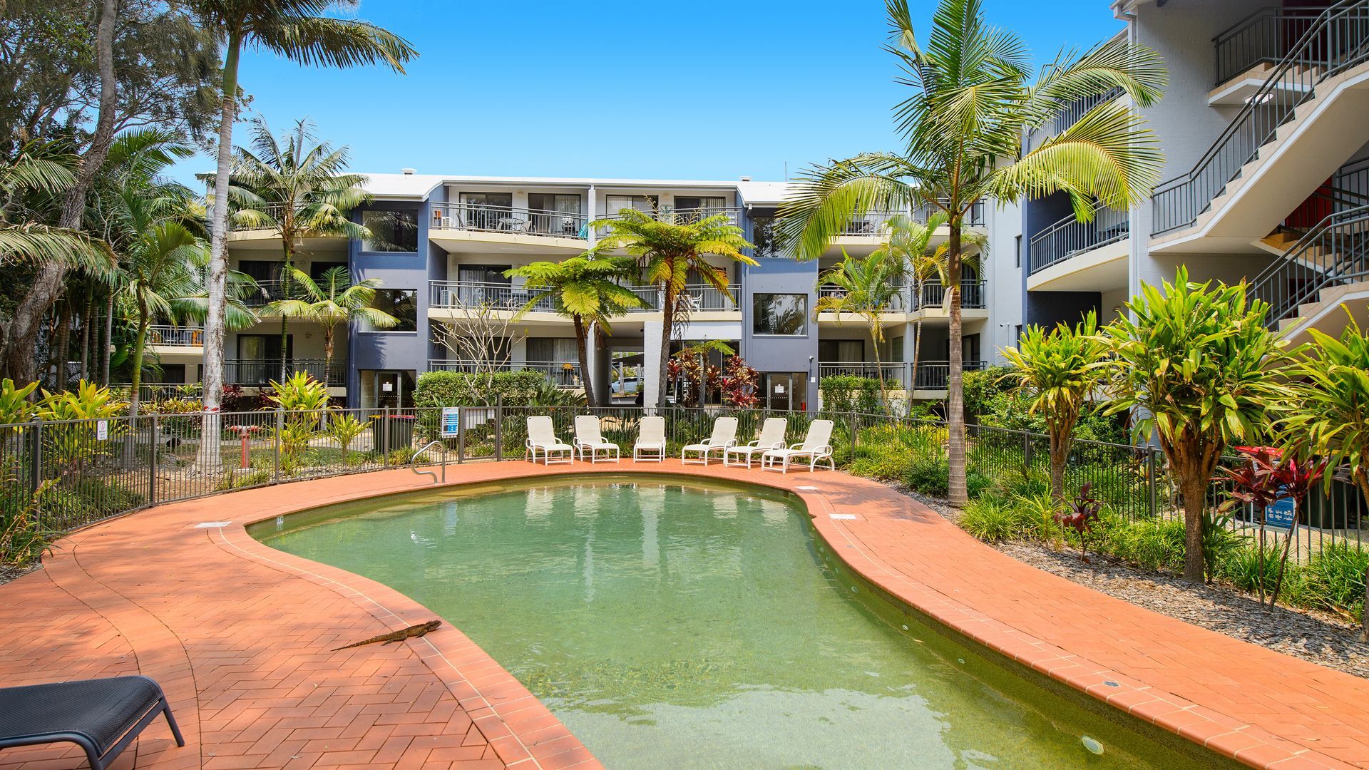 A pool area with lounge chairs, a brick pathway, and a multi-story building with balconies.