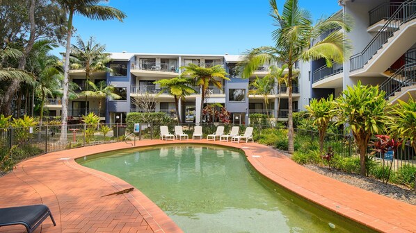 A pool area with lounge chairs, a brick pathway, and a multi-story building with balconies.
