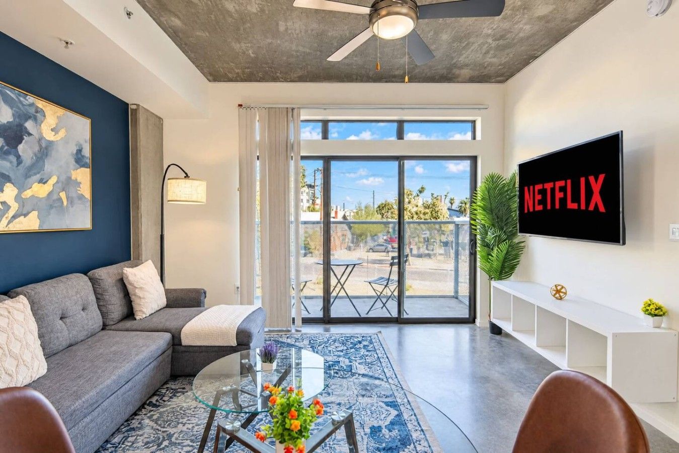 Ein modernes Wohnzimmer mit einem grauen Sofa, einem Glastisch mit Blumen, einem Deckenventilator und einem großen Fenster mit Ausblick.