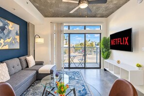A modern living room with a grey sofa, a glass coffee table with flowers, a ceiling fan, and a large window with a view of the outside.