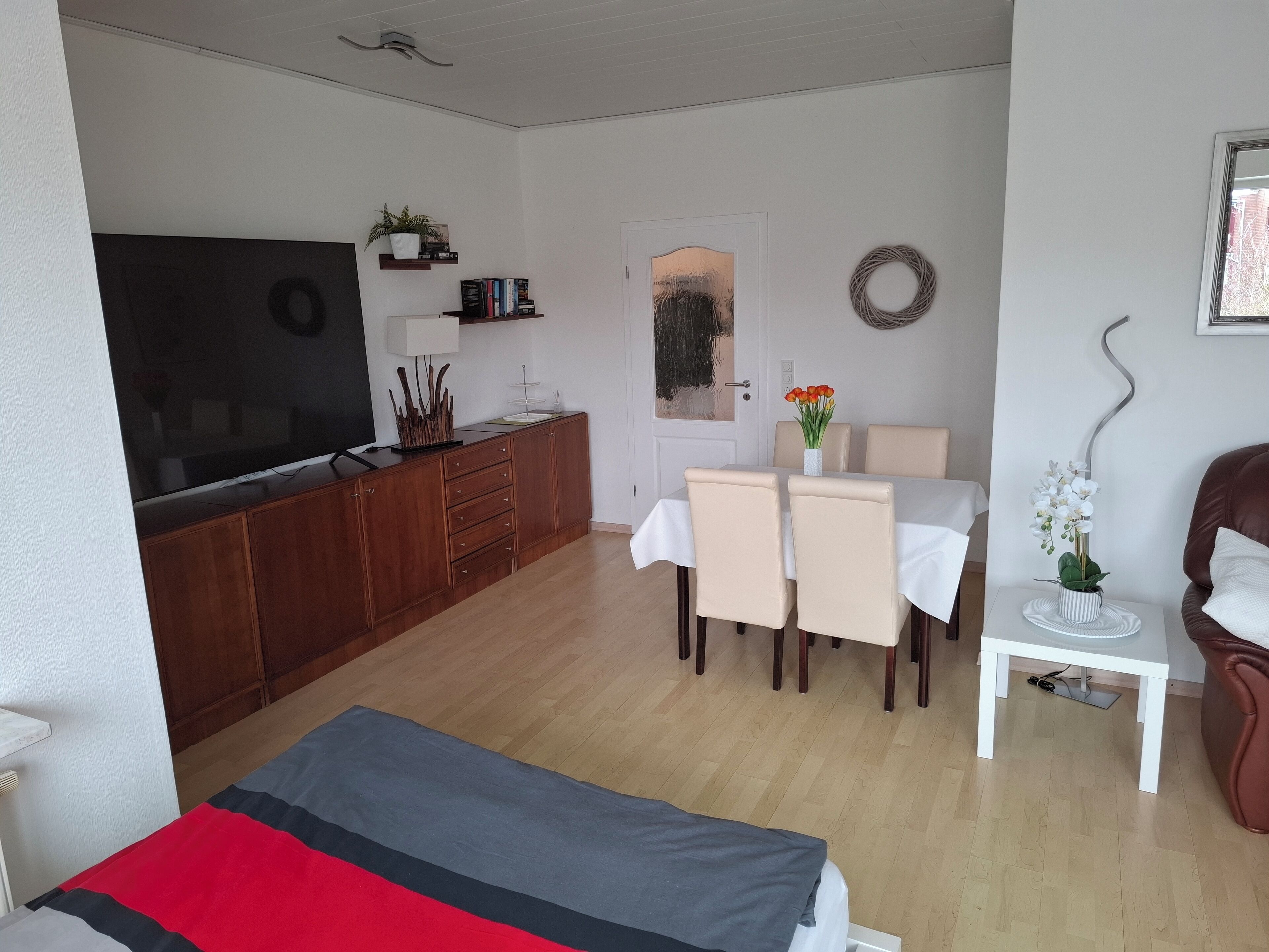 A modern living room with a flat-screen TV, wooden cabinets, a dining table with chairs, and a small white table with a vase of flowers.