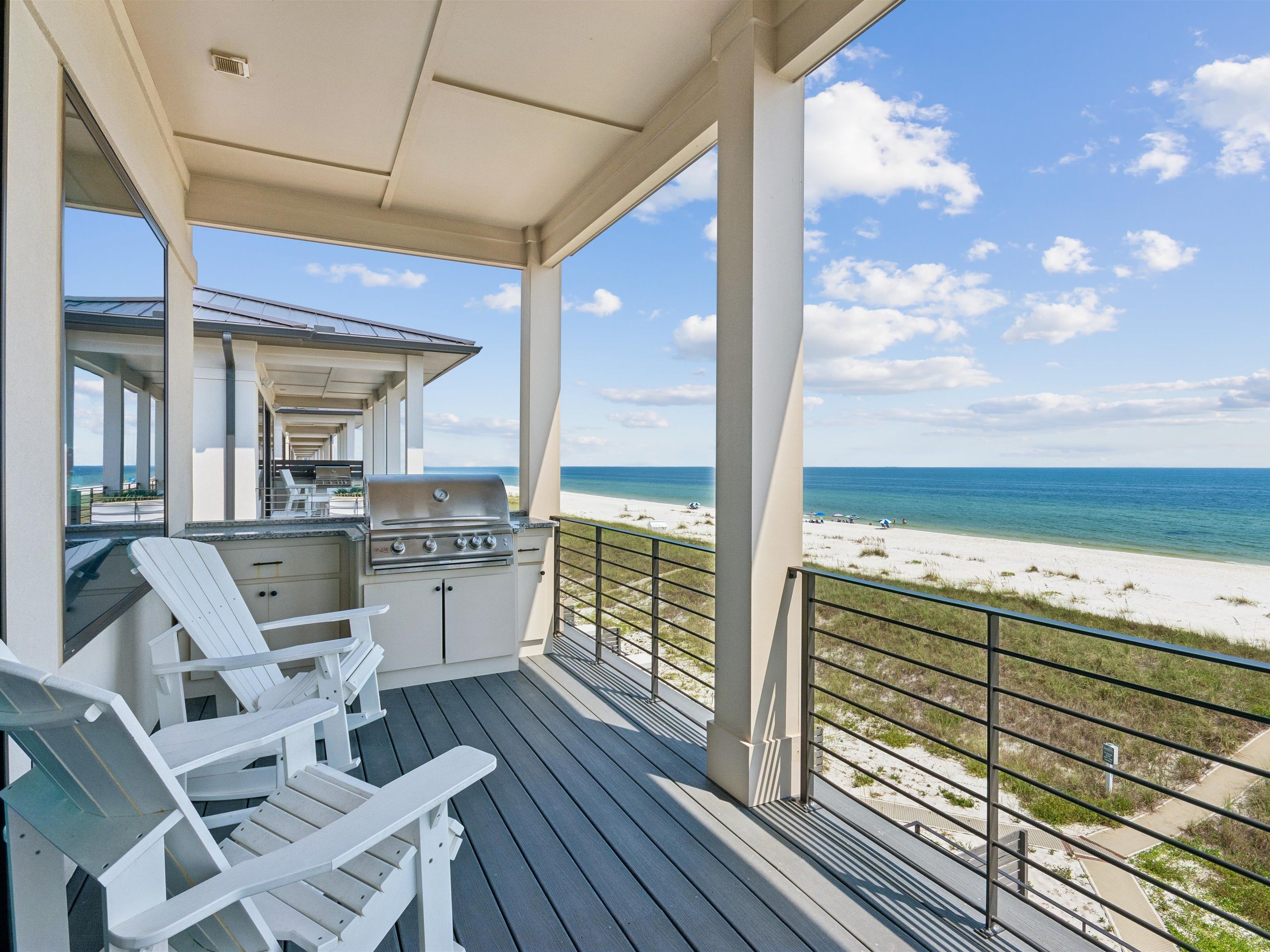 A covered outdoor area with a grill, white Adirondack chairs, and a view of the beach.