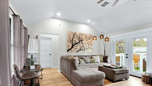 A modern living room with a grey sofa, a dining table with chairs, a large window, and a wall-mounted artwork. - Little White House of Wimberley I River access (Wimberley)