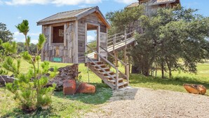 Une cabane en bois rustique, dotée d’un escalier menant à une mezzanine, entourée de verdure et d’arbres.
