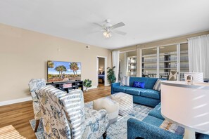 Un salon comprenant un canapé, un fauteuil, un pouf, une télévision, un ventilateur de plafond et donnant sur un balcon.