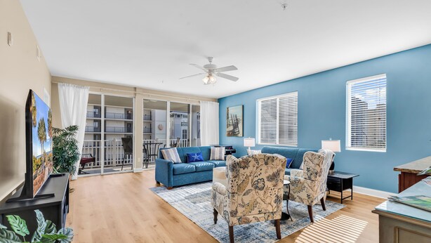 A living room with a blue sofa, patterned armchairs, a ceiling fan, and a view through a glass door to an outdoor balcony. - Ocean View l 5BR l Pool l Sleeps 16 l Luxury Villa (north myrtle beach)