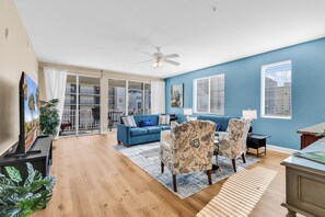 Ein Wohnzimmer mit blauem Sofa, gemusterten Sesseln, einem Deckenventilator und Blick durch eine Glastür auf einen Außenbalkon.