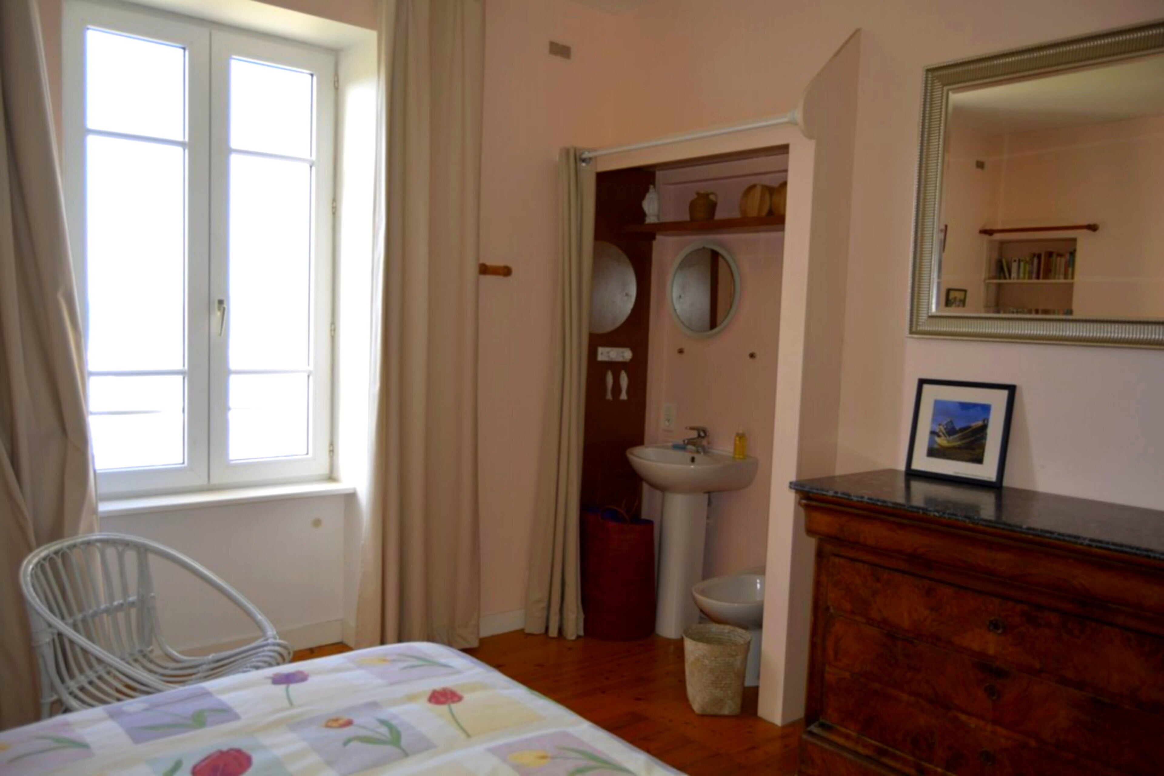 Une chambre à coucher comprenant un lit, une chaise, une commode, un miroir, une fenêtre et une salle de bain avec un lavabo et des toilettes.
