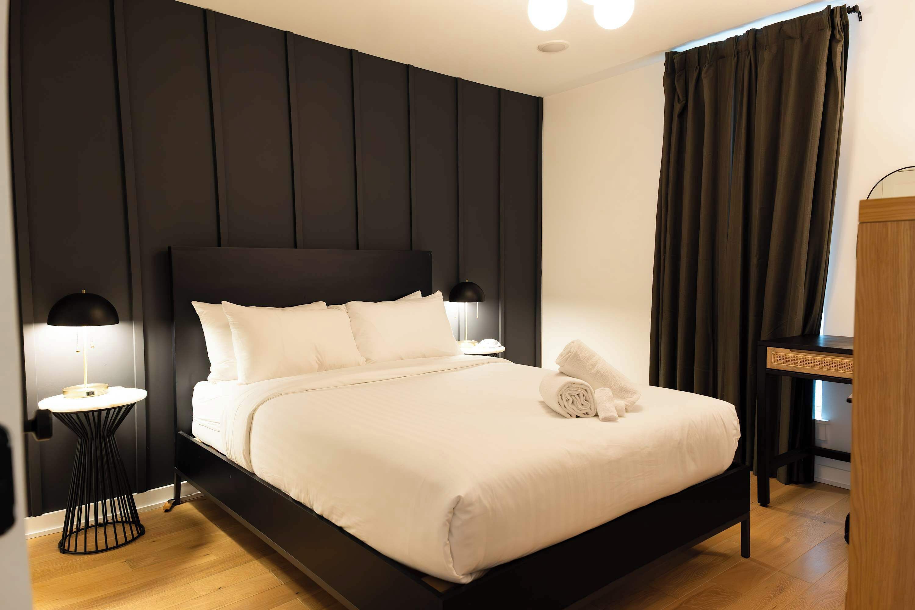 Een netjes opgemaakt bed met witte lakens, een zwart hoofdeinde en een klein rond tafeltje met een lamp ernaast.