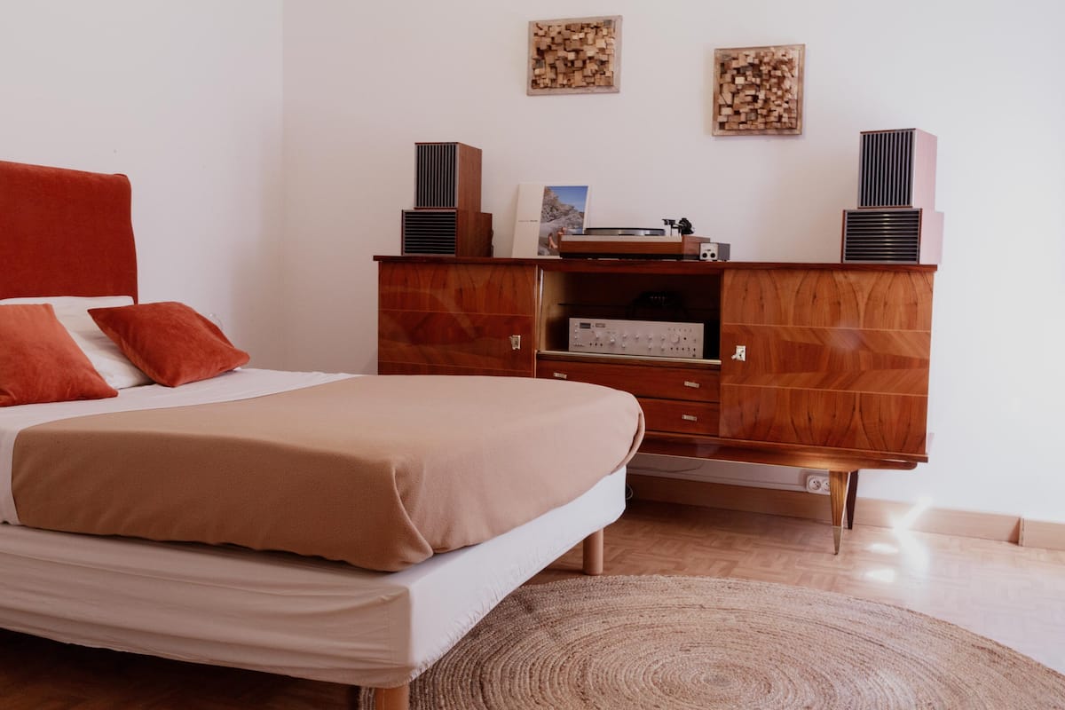Ein Schlafzimmer mit einem Bett, einem Holzschrank, einem kleinen Tisch und zwei Lautsprechern an der Wand.