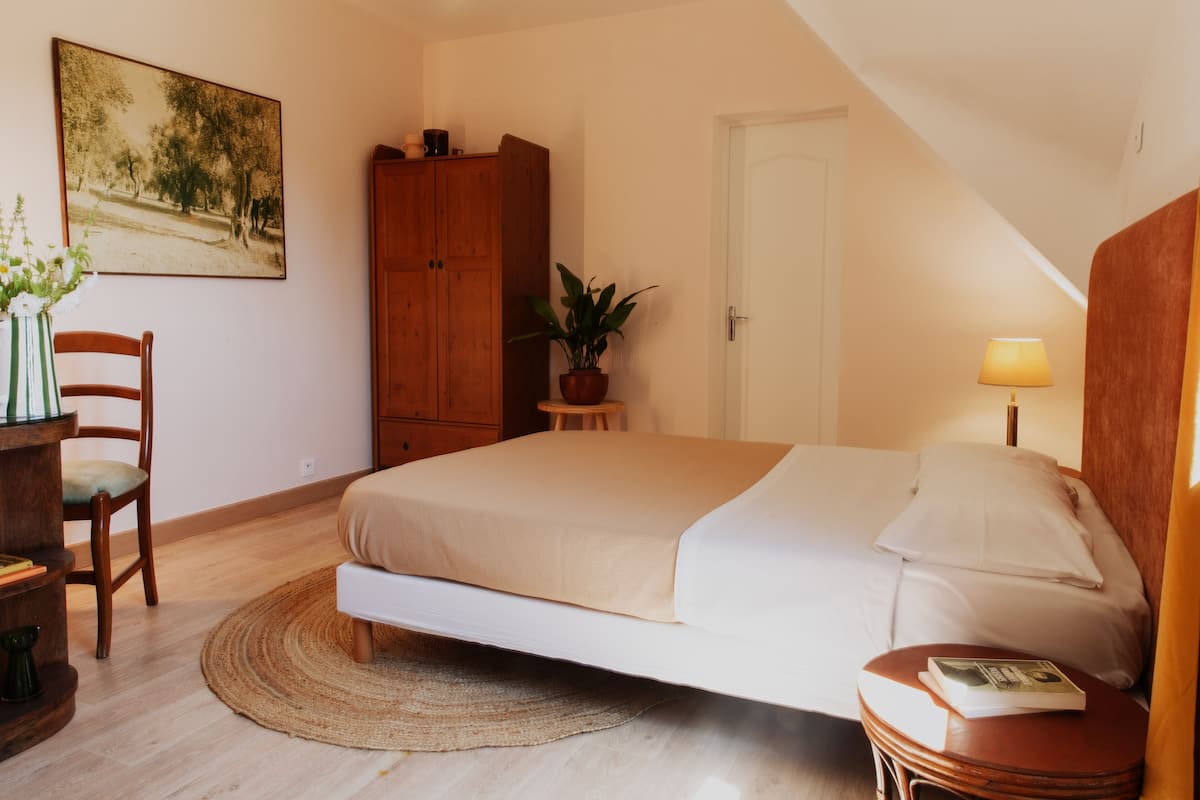Ein Schlafzimmer mit Bett, Schreibtisch mit Stuhl, einem Holzschrank, einer Topfpflanze und einer Tür.