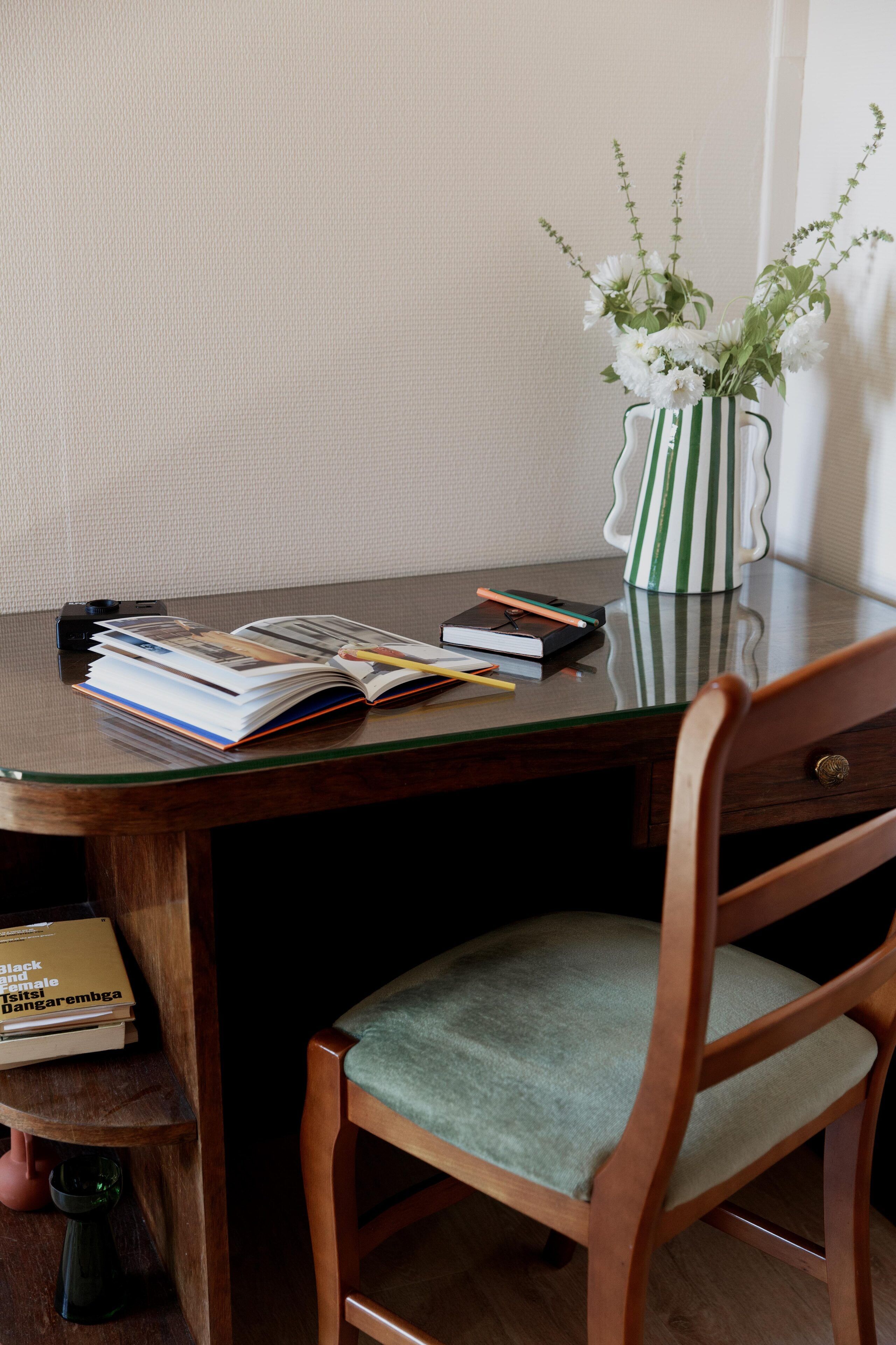 A wooden desk with a glass top, a vase with white flowers, and a chair with a cushioned seat.