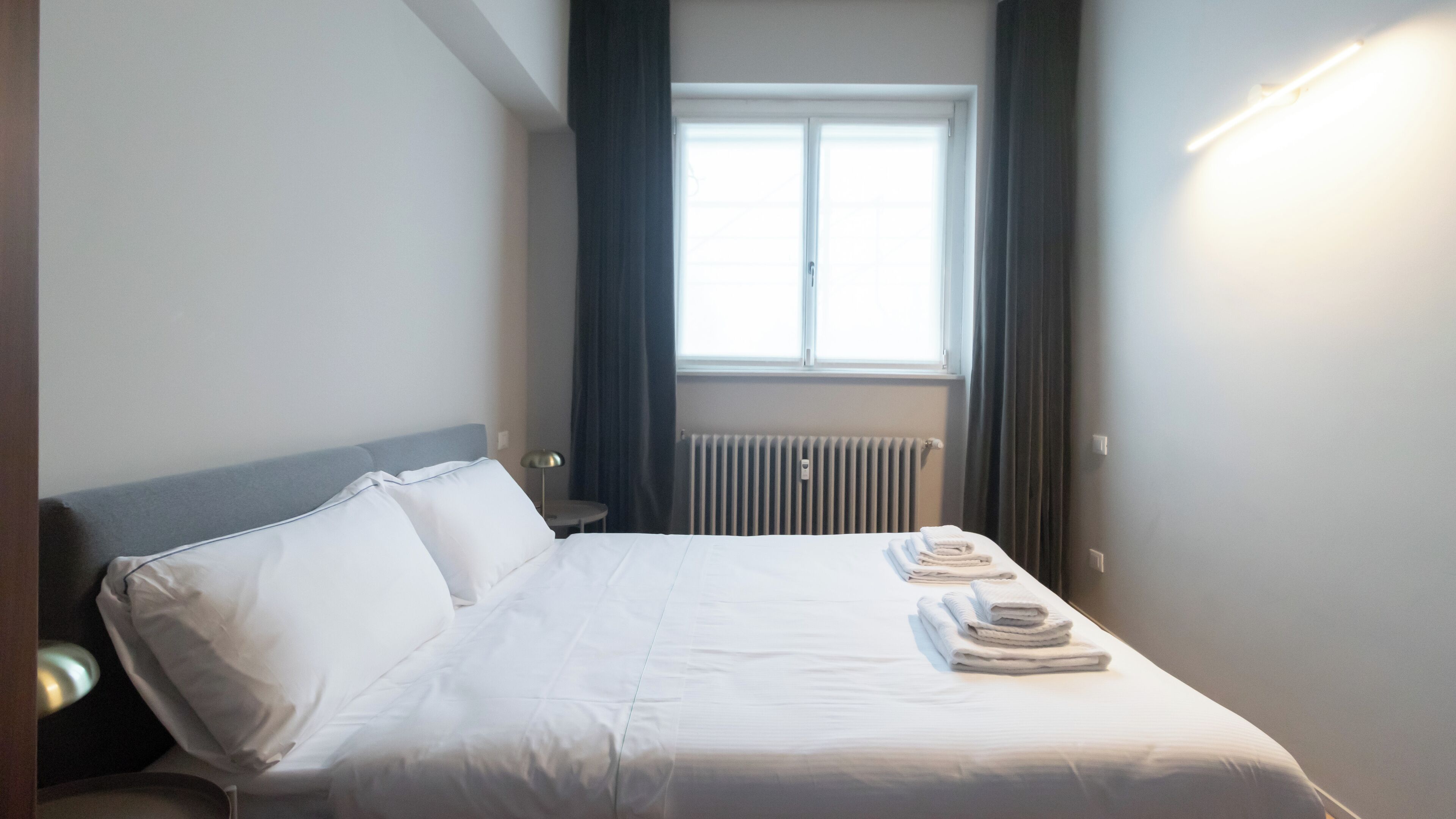 A neatly made bed with white linens, a bedside table with a lamp, and a window with curtains.
