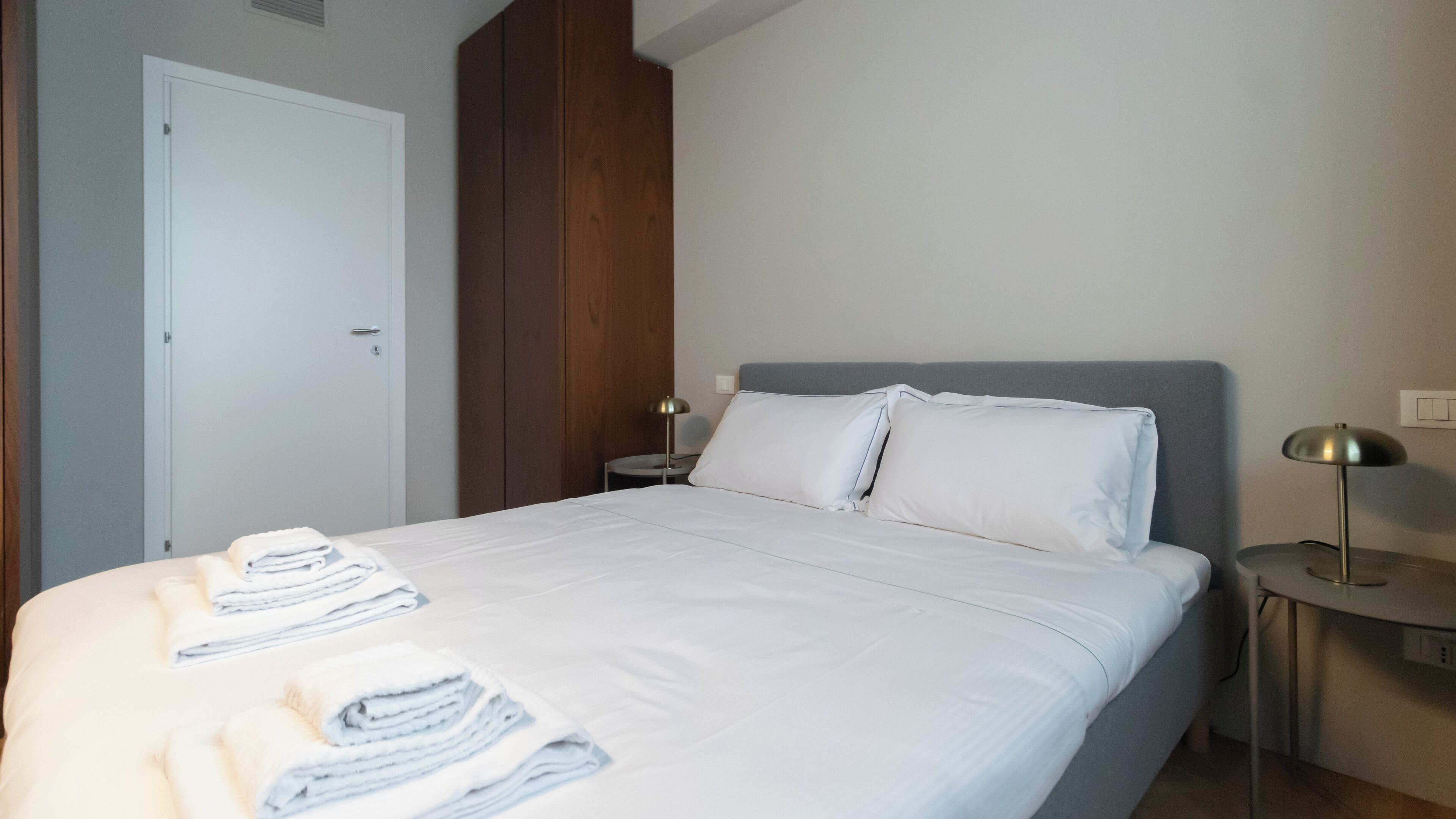 A neatly made bed with white linens and folded towels, a bedside lamp, and a wooden wardrobe in a hotel room.
